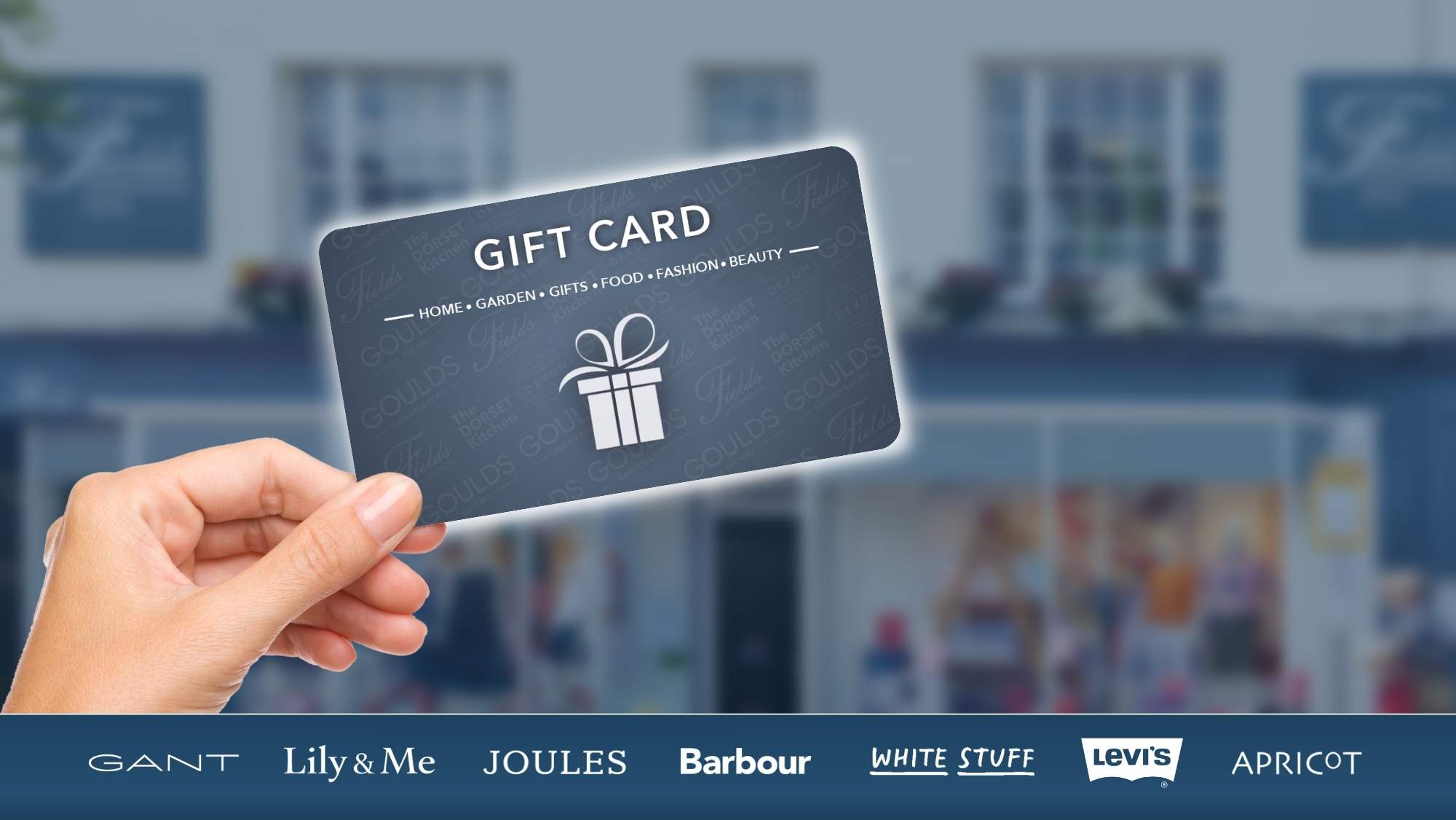 A hand holding a Fields Of Sidmouth gift card, in front of the store, a brand bar with a variety of fashion brand logos