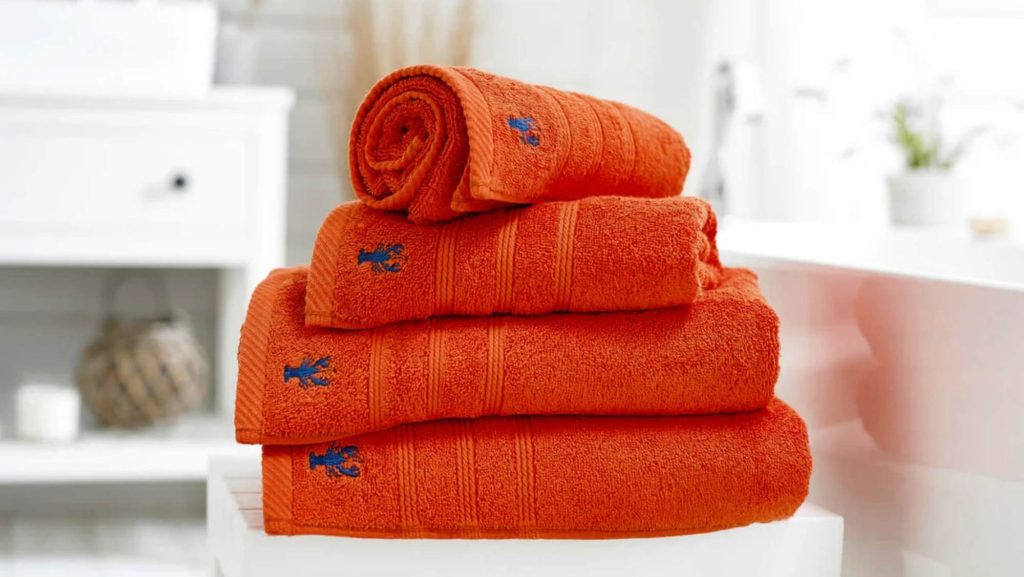 A stack of vibrant orange bath towels in various sizes (face cloth, hand towel, bath towel, bath sheet, bath mat) with an embroidered blue lobster.