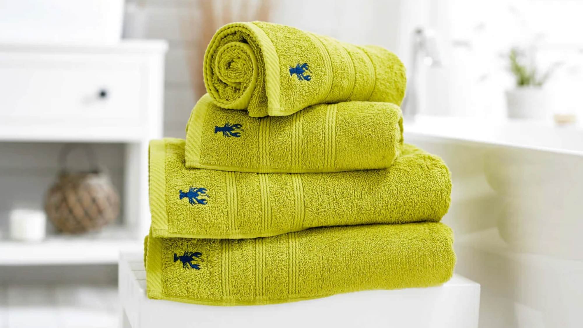 A stack of lime green bath towels in various sizes (face cloth, hand towel, bath towel, bath sheet, bath mat) with an embroidered blue lobster. Available at Fields Of Sidmouth