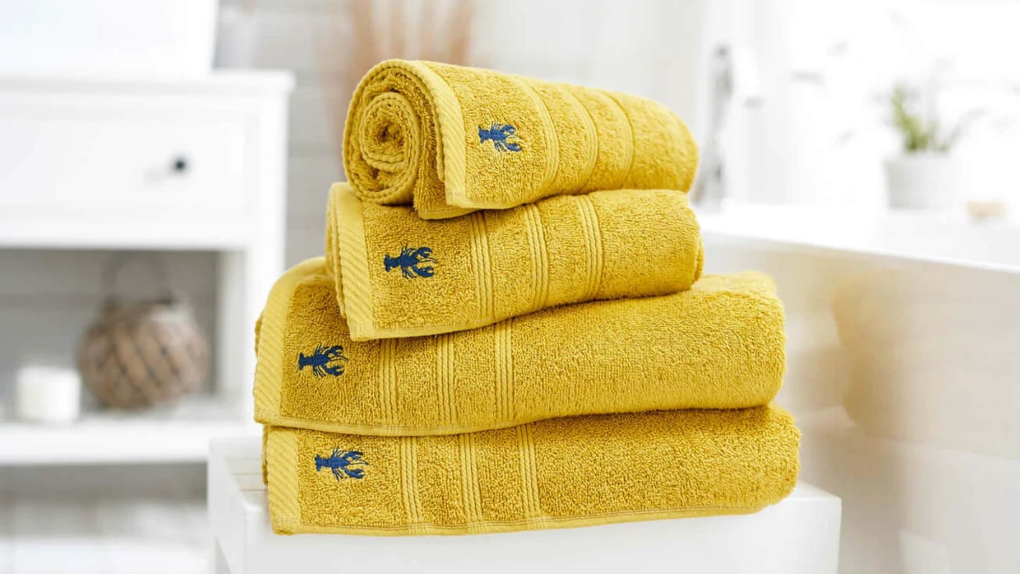 A stack of bright yellow bath towels in various sizes (face cloth, hand towel, bath towel, bath sheet, bath mat) with an embroidered blue lobster. Available at Fields Of Sidmouth